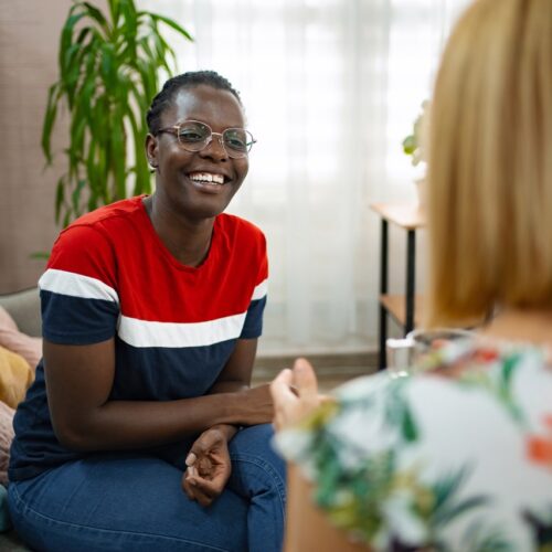 In a cozy living room filled with plants, two friends engage in a joyful conversation. One woman, wearing a colorful striped shirt, smiles warmly while listening attentively to her companion.