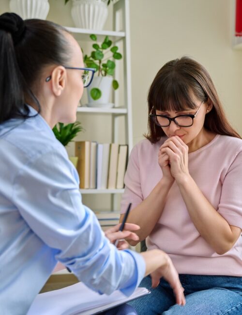 Mature sad woman in therapy session with mental professional psychologist, sitting at table in office of therapist counselor social worker. Psychology psychotherapy mental assistance treatment support