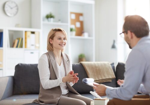 Young blonde smiling woman sitting on couch and looking at counselor during discussion of ways of solving problem